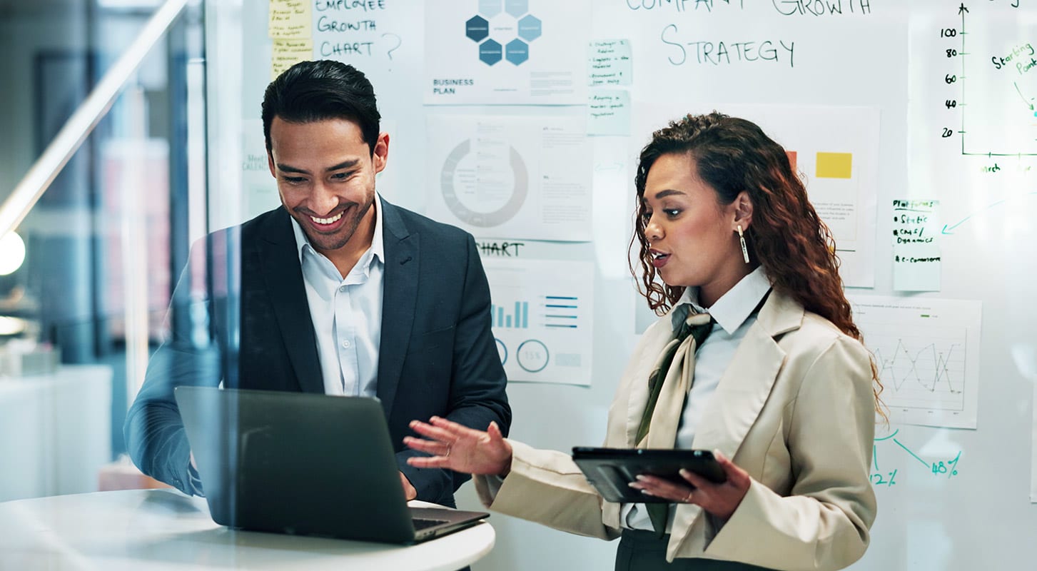 deux professionnels collaborent debout autour d'une table, examinant des données sur un ordinateur portable et une tablette dans un bureau moderne, avec des tableaux, des graphiques et des notes stratégiques affichés sur un tableau blanc derrière eux.